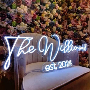 May include: White neon sign with the text "The Williams est. 2024" on a white sofa in front of a wall of pink and purple flowers.