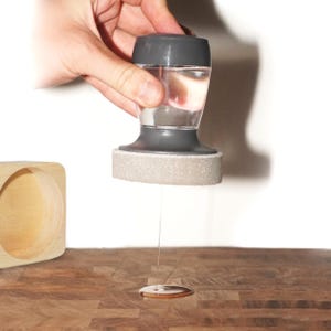 May include: A gray and clear plastic dispenser with a gray sponge on the bottom. The dispenser is being squeezed and a clear liquid is dripping onto a wooden surface.