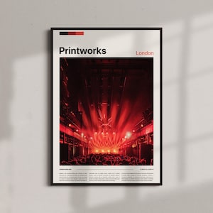 May include: Framed poster featuring the Printworks London logo with a red and black color scheme. The image inside the frame showcases a concert with bright red stage lights illuminating a crowd of people. The poster is mounted on a white wall.