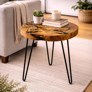 May include: A round wooden side table with a unique, natural wood grain pattern. The table has black hairpin legs and is topped with a small potted plant, books, and a candle. The table is approximately 20 inches in diameter.