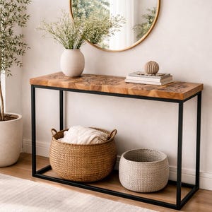 May include: A modern entryway table with a black metal frame and a natural wood top. Two woven baskets sit below, one filled with a blanket. A round gold mirror and a vase of flowers are on top, creating a stylish decor.