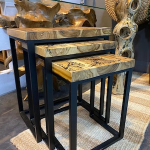 Puede incluir: Un juego de tres mesas nido con tapas de madera natural y marcos de metal negro. Los patrones de vetas de la madera varían, mostrando diseños únicos. Las mesas son de diferentes tamaños, diseñadas para encajar una debajo de la otra. La estética general es moderna y rústica.