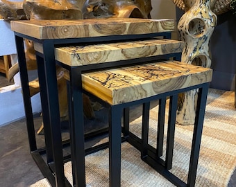 May include: A set of three nesting tables with natural wood tops and black metal frames. The wood grain patterns vary, showcasing unique designs. The tables are of different sizes, designed to fit one under the other. The overall aesthetic is modern and rustic.