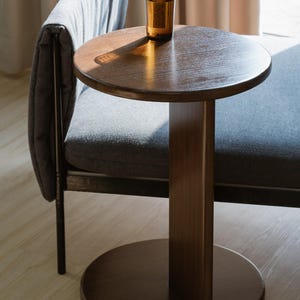 May include: A small, round wooden side table with a single central support and a circular base. A glass of amber liquid sits on the table's surface. The table is next to a gray upholstered bench.