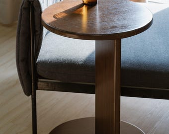 May include: A small, round wooden side table with a single central support and a circular base. A glass of amber liquid sits on the table's surface. The table is next to a gray upholstered bench.