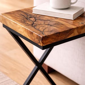 May include: A small, square side table with a unique wood grain top and black metal legs. The tabletop features a striking, natural pattern. A white book and a ceramic mug sit on top of the table. The table is approximately 20 inches wide.