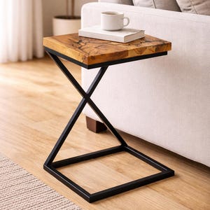 May include: A square side table with a natural wood top and a black metal frame. The tabletop has a unique wood grain pattern. A book and a white mug sit on top of the table. The table is next to a white sofa.