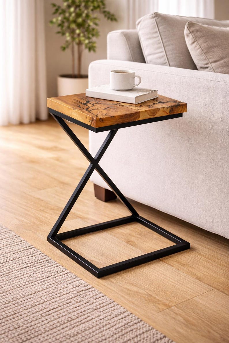 May include: A square side table with a natural wood top and a black metal frame. The tabletop has a unique wood grain pattern. A book and a white mug sit on top of the table. The table is next to a white sofa.