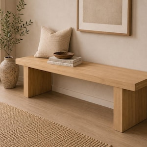 May include: A light oak bench with a rectangular top and two square legs. A beige cushion, a wooden bowl, and two books are on the bench. A decorative vase with a plant is to the left of the bench.
