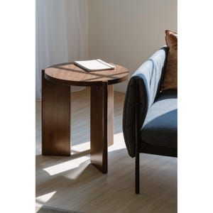 May include: A dark brown wooden side table with a round top and two rectangular legs. A white notebook rests on the table's surface. The table is next to a blue sofa with a brown pillow.
