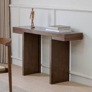 May include: A dark brown wooden console table with a rectangular top and two vertical supports. Two books and a small wooden artist's mannequin are on the table. The table is against a white wall.