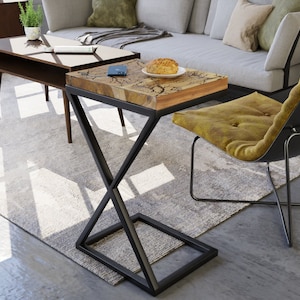 Puede incluir: Una pequeña mesa auxiliar de madera con una base en forma de X de metal negro. La mesa tiene un acabado de madera natural y está sobre una alfombra gris claro. Un sofá gris y una silla amarilla están en el fondo.