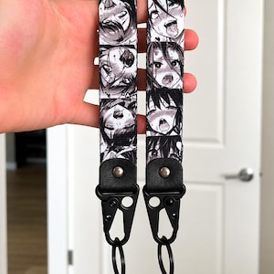 Puede incluir: Dos cordones estampados en blanco y negro con caras de estilo anime. Cada cordón tiene una pieza final de cuero negro con un cierre de metal y un anillo de metal. Los cordones se sostienen sobre un fondo blanco.