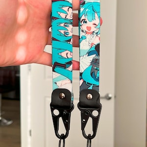 Op de afbeelding: Twee turquoise en witte lanyards met een cartoonkarakterontwerp. Elke lanyard heeft een zwarte metalen sluiting en een zwart lederen accent. Het personage heeft turquoise haar en draagt een witte outfit.