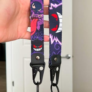 May include: Two purple and black keychains with cartoon character designs. One features a character with a long red tongue, the other with a white and red design. Each keychain has a black metal clasp and a key ring.