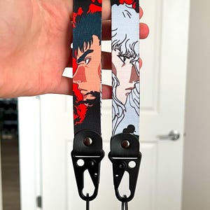 Può includere: Due cordini neri con ritratti di personaggi anime. Uno presenta un design rosso e nero con un profilo laterale di un personaggio. L'altro ha un design azzurro e bianco con un ritratto di un personaggio. Ogni cordino ha un fermaglio in metallo nero.