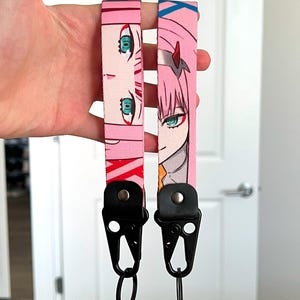May include: Two pink keychains with anime-style character illustrations. Each keychain has a black clasp and a metal ring. The character art features pink hair, green eyes, and a red accent.