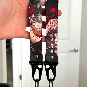 Op de afbeelding: Twee zwarte lanyards met anime-personageafbeeldingen. Elke lanyard heeft een zwarte metalen sluiting en een splitring. De afbeeldingen zijn van een personage met zwart haar, rode ogen en een rood-zwart ontwerp.