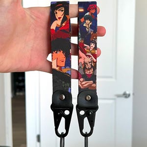 May include: Two black lanyards with colourful anime character illustrations. Each lanyard has a black metal clip and a key ring. The lanyards are held up against a white door.