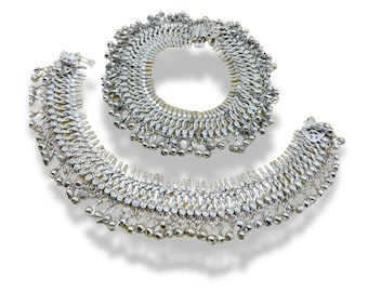 May include: A pair of silver anklets with intricate designs and dangling bells. The anklets are made of small, interconnected metal pieces that create a delicate and ornate look.