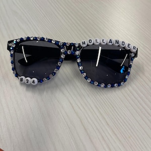 May include: Black sunglasses with dark lenses, embellished with blue and clear rhinestones around the frames. The word "HOLLANDER" is spelled out in white letter beads across the top of the left lens, and the number "24" is on the left side.