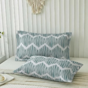 Puede incluir: Dos fundas de almohada rectangulares con un patrón geométrico en verde azulado y blanco, compuesto por líneas verticales dispuestas en una formación de zigzag repetitiva. La tela muestra un estampado consistente en ambas fundas horizontales.