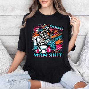 May include: A black t-shirt with a colorful graphic of a skeleton sitting on a toilet, holding a phone and wearing headphones. The text "Doing Mom Shit" is printed below the graphic.