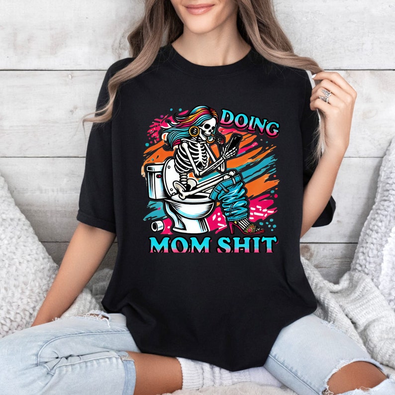 May include: A black t-shirt with a colorful graphic of a skeleton sitting on a toilet, holding a phone and wearing headphones. The text "Doing Mom Shit" is printed below the graphic.