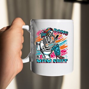May include: White ceramic coffee mug with a colorful illustration of a skeleton sitting on a toilet and holding a phone. The text "Doing Mom Shit" is printed on the mug.