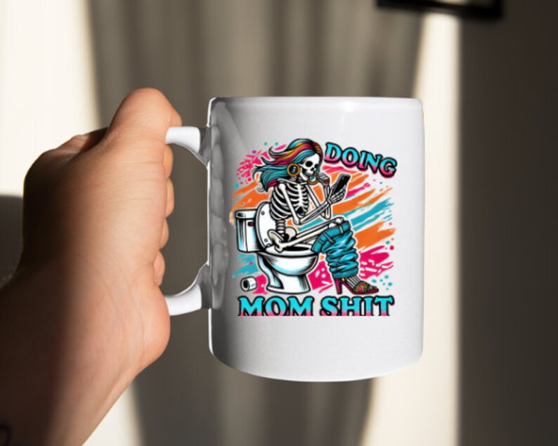 May include: White ceramic coffee mug with a colorful illustration of a skeleton sitting on a toilet and holding a phone. The text "Doing Mom Shit" is printed on the mug.