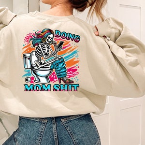 May include: A beige sweatshirt with a colorful graphic of a skeleton sitting on a toilet and holding a phone. The text "Doing Mom Shit" is printed below the graphic.