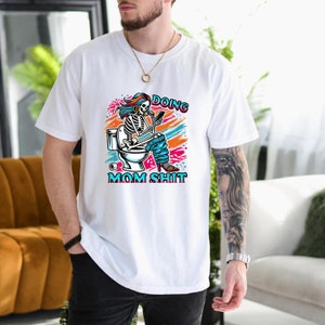 May include: White t-shirt with a colorful graphic of a skeleton sitting on a toilet with the text "Doing Mom Shit".