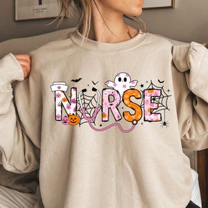 May include: Beige sweatshirt with the word "NURSE" in colorful Halloween-themed lettering. The design includes a nurse's hat, pumpkins, a ghost, spiderwebs, bats, a stethoscope, and spiders. A festive and fun design.