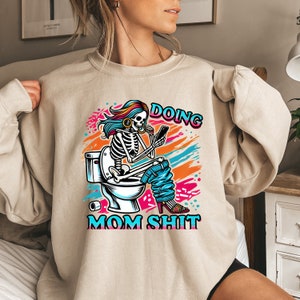 May include: A beige sweatshirt with a graphic print of a skeleton sitting on a toilet, holding a phone and a cup. The text "Doing Mom Shit" is printed below the image.