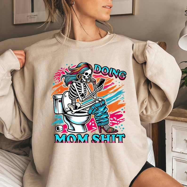 May include: A beige sweatshirt with a graphic print of a skeleton sitting on a toilet, holding a phone and a cup. The text "Doing Mom Shit" is printed below the image.