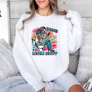 May include: White sweatshirt with a colorful graphic of a skeleton sitting on a toilet, holding a phone and wearing a blue jean and a pink high heel. The text "Doing Mom Shit" is printed below the graphic.