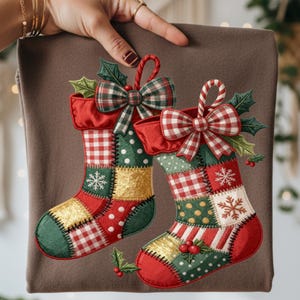 May include: A brown Christmas-themed decorative cushion featuring two embroidered stockings. The stockings are patchwork with red, green, and gold fabrics, adorned with bows, holly, and snowflakes. The cushion is held by a person.