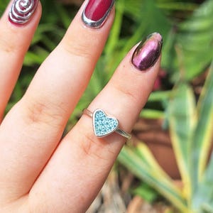 May include: A silver ring featuring a heart-shaped turquoise stone. The ring is worn on a finger with manicured nails. The heart is filled with small, light blue dots, creating a textured look. The ring band is thin and delicate.