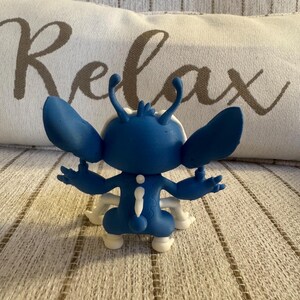 K&ouml;nnte beinhalten: Blaue Figur einer Zeichentrickfigur mit gro&szlig;en Ohren und wei&szlig;en Akzenten. Die Figur steht auf einer gemusterten Oberfl&auml;che. Das Wort "Relax" ist auf einem Kissen im Hintergrund zu sehen.