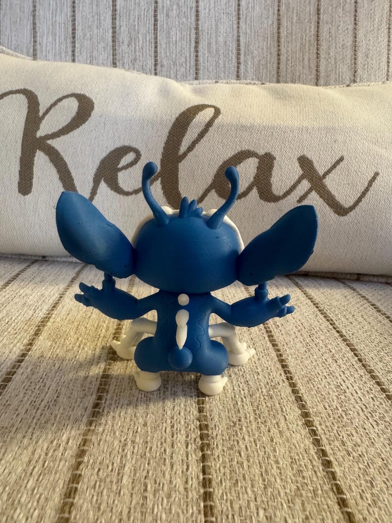 K&ouml;nnte beinhalten: Blaue Figur einer Zeichentrickfigur mit gro&szlig;en Ohren und wei&szlig;en Akzenten. Die Figur steht auf einer gemusterten Oberfl&auml;che. Das Wort "Relax" ist auf einem Kissen im Hintergrund zu sehen.