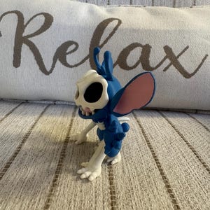 K&ouml;nnte beinhalten: Eine blau-wei&szlig;e Figur eines Charakters mit gro&szlig;en Ohren und einem sch&auml;delartigen Gesicht. Die Figur steht auf einer strukturierten Oberfl&auml;che, im Hintergrund ein Kissen mit der Aufschrift "Relax" in Schreibschrift.