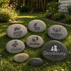 May include: A selection of engraved memorial stones for pets, featuring names and dates. The stones display various designs, including a turtle, bird, snake, paw print, and cat and dog silhouettes. Names include Speedy, Kiwi, Charlie, Slither, Jordan, Max, Milo, and Ozzy & Dutty. The stones are on grass.