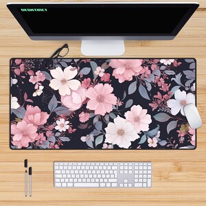 May include: A black desk mat with a floral pattern featuring pink and white flowers on a dark background. The mat is designed for use with a computer mouse and keyboard.