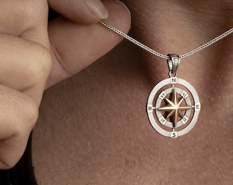 May include: A silver chain necklace with a compass pendant. The compass is silver with a gold-coloured star in the centre. The compass face has the letters N, S, E, and W around the edge.