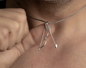 May include: A silver necklace featuring a bow and arrow pendant. The pendant is a detailed miniature of a bow and arrow, with the arrow pointing downwards. The necklace is worn around the neck.