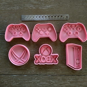 May include: Six pink cookie cutters in the shape of Xbox controllers, the Xbox logo, and a console. A metal ruler is placed above the cookie cutters. The cookie cutters are made of plastic.