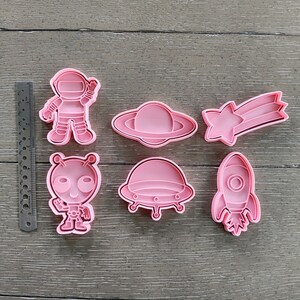 May include: Six pink plastic cookie cutters shaped like an astronaut, alien, spaceship, shooting star, and two different types of flying saucers.
