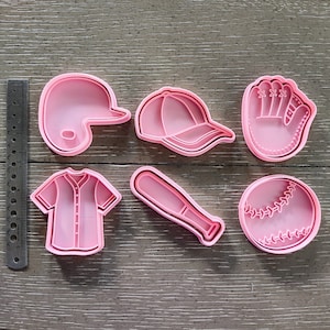 May include: Six pink plastic cookie cutters shaped like a baseball, baseball bat, baseball glove, baseball cap, baseball jersey, and a baseball.