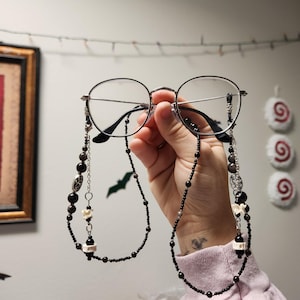 May include: Black beaded glasses chain with silver accents and small white charms. The chain is made of small black beads and silver beads. The charms are in the shape of bats and other small shapes.