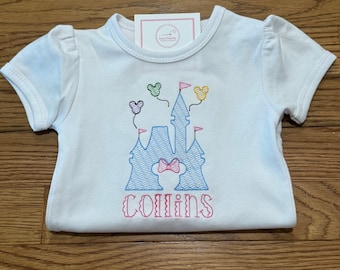 May include: A white short-sleeved shirt featuring an embroidered blue castle outline with a pink bow silhouette, colorful floating balloons, and the name Collins in pink lettering. A small white tag with the text Fancy Mommy is at the neckline.